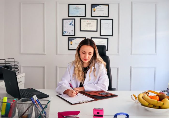 Nutritionist sits at a modern office desk writing, with fruits and diplomas displayed, emphasizing health and professionalism.